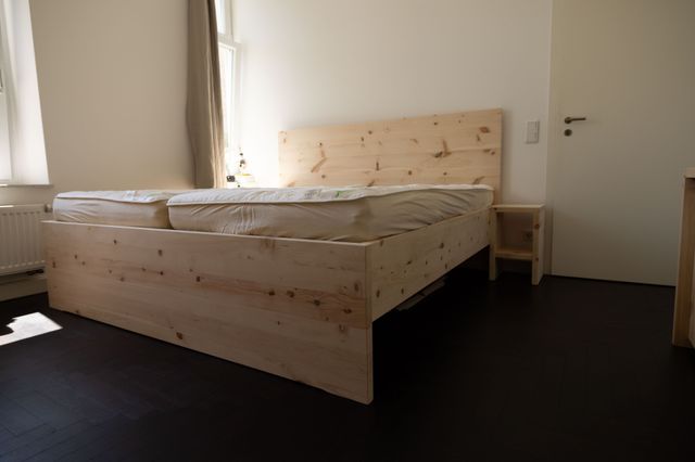 Massives Doppelbett in Zirbe. Das Fußteil geht bis auf den Boden. Das Kopfteil ist grade. Daneben steht ein Nachttisch, ohne Schublade. 