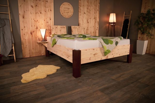 Zirbenbett mit dunklen Nussbaumfüßen. Auf dem Boden ist ein kleines Fell. Die Wand ist teilweise mit Holz verkleidet. 