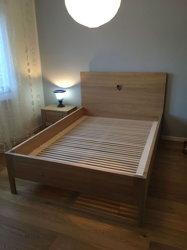 Eiche Massivholzbett. Im Kopfteil ist ein Herz eingefräst. Neben dem Bett ist ein Nachttisch, mit einer Schublade.
