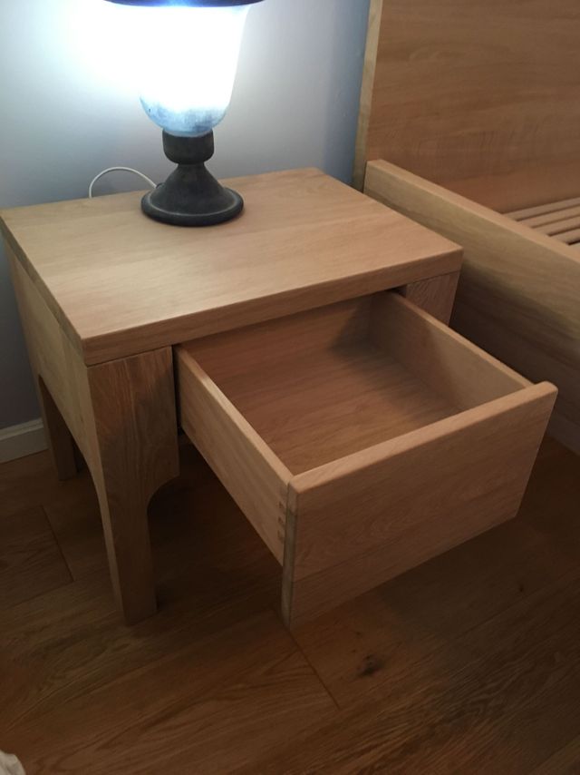 Nachtkästchen, mit einer Holzschublade. Das Möbel ist in warmer Eiche gefertigt. Auf dem Nachttisch steht eine Lampe. 
